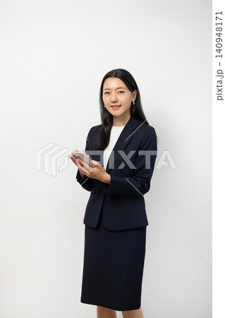白い壁の前でスマートフォンを持ってカメラ目線で微笑む若い日本人のビジネスウーマン 白い壁の前でスマートフォンを持ってカメラ目線で微笑む若い日本人のビジネスウーマン 140948171