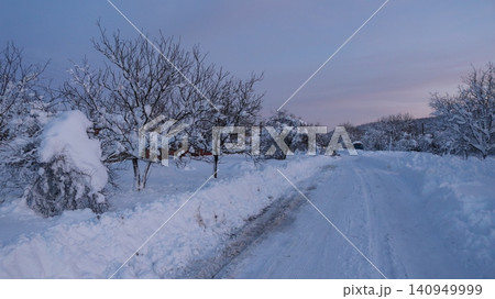 A cleared country road in the winter twilight. High snowdrifts line the roadsides, and the trees and bushes are thickly covered with a fluffy crust. The sky on the horizon is painted in soft pink tone A cleared country road in the winter twilight. High snowdrifts line the roadsides, and the trees and bushes are thickly covered with a fluffy crust. The sky on the horizon is painted in soft pink tone 140949999