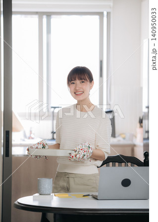 Asian woman kitchen cupcake tray smile home baking laptop table, bright window, white sweater, cheerful expression, homemade dessert preparation in clean modern room 140950033