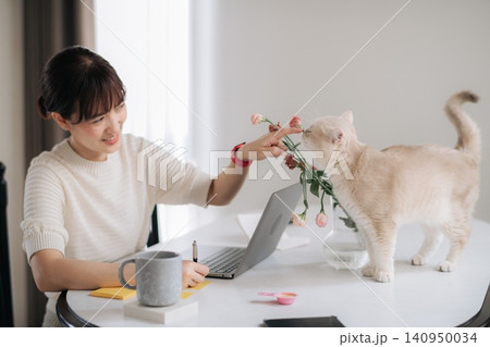 Asian woman cat laptop flower table home indoor smile white sweater at round desk, gentle pet moment with bouquet, mug, notebook, pen, soft daylight near window 140950034