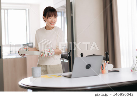 Asian woman, kitchen, laptop, cooking, oven mitt, home office, smiling expression while preparing food beside table in bright apartment workspace with cup and stationery Asian woman, kitchen, laptop, cooking, oven mitt, home office, smiling expression while preparing food beside table in bright apartment workspace with cup and stationery 140950042