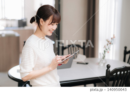 Asian woman tablet kitchen home smile, young adult with relaxed expression using digital device near dining table, bright window, flower vase, chair, soft indoor light 140950043