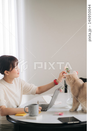 Asian woman cat laptop home office flower on desk, calm remote work moment by window with pet sniffing blossom, mug, notebook, phone, soft light, cozy workspace lifestyle 140950044
