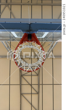 A vertical low-angle shot looking directly up from underneath a red basketball hoop and net, showcasing the symmetrical geometric steel roof structure of an indoor gym stadium. 140951021