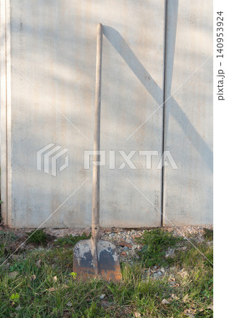 A lone shovel rests against a textured concrete wall on a bright sunny day. The contrast between the rough concrete surface and the metal tool highlights an industrial and outdoor work setting 140953924