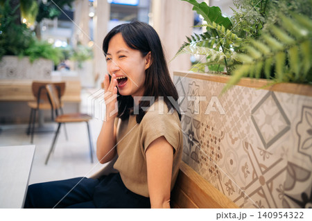 Smiling woman indoor casual clothing sitting wooden bench patterned tile green plant cheerful candid Asian relaxed modern cafe natural light 140954322