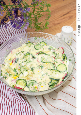 A bowl of okroshka filled with potatoes, chicken breast, cucumbers, herbs, and boiled eggs sits on a table. Nearby, a floral vase adds color to the setting A bowl of okroshka filled with potatoes, chicken breast, cucumbers, herbs, and boiled eggs sits on a table. Nearby, a floral vase adds color to the setting 140954937