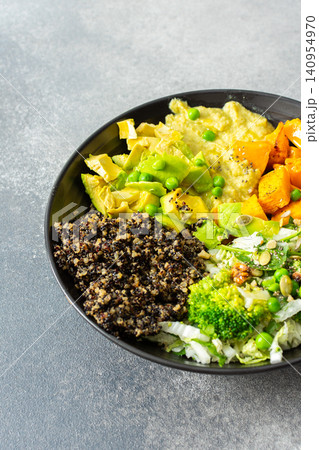 A quinoa bowl is arranged with roasted pumpkin, hummus, and a fresh salad. Plates are set on a table with utensils. Sunshine comes through the window 140954970