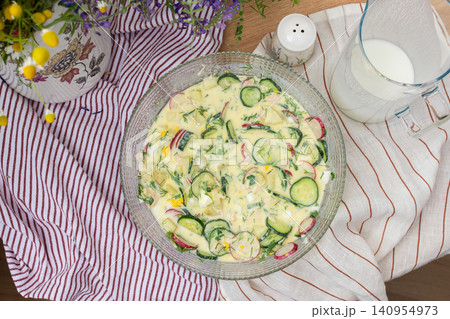 A bowl of okroshka filled with potatoes, chicken breast, cucumbers, herbs, and boiled eggs sits on a table. Nearby, a floral vase adds color to the setting A bowl of okroshka filled with potatoes, chicken breast, cucumbers, herbs, and boiled eggs sits on a table. Nearby, a floral vase adds color to the setting 140954973