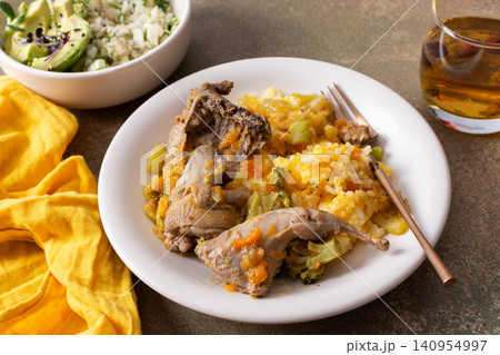 Rice mixed with stewed quail is served on a plate alongside fresh salad and avocado. A drink is present, creating a balanced meal perfect for breakfast or lunch 140954997