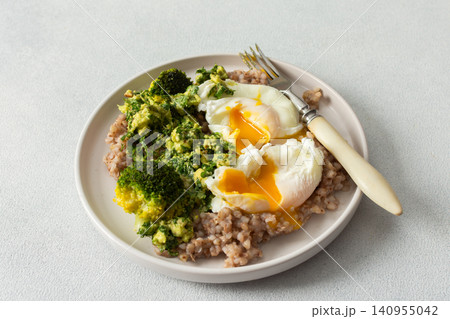 A healthy breakfast bowl features green buckwheat topped with pesto, steamed broccoli, and poached eggs. A fork rests beside the dish ready for a nutritious meal A healthy breakfast bowl features green buckwheat topped with pesto, steamed broccoli, and poached eggs. A fork rests beside the dish ready for a nutritious meal 140955042