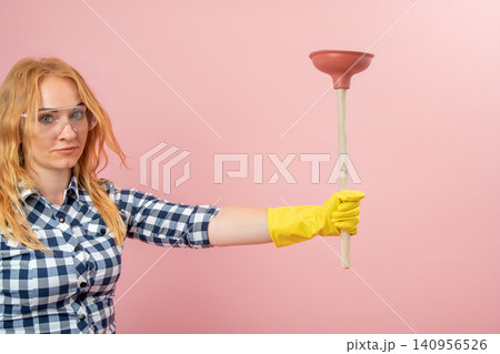 Young woman wearing a plaid shirt and protective eyewear with a serious expression, holding a toilet plunger, representing concepts of plumbing, cleaning, and DIY home repair Young woman wearing a plaid shirt and protective eyewear with a serious expression, holding a toilet plunger, representing concepts of plumbing, cleaning, and DIY home repair 140956526