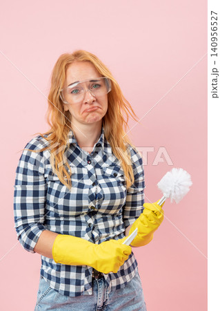 Woman wearing protective eyewear and yellow gloves holding a toilet brush, showing a face of deep displeasure and unwillingness to undertake household chores Woman wearing protective eyewear and yellow gloves holding a toilet brush, showing a face of deep displeasure and unwillingness to undertake household chores 140956527