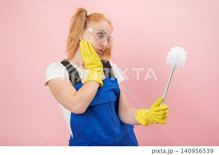 Woman wearing blue overalls, yellow gloves, and safety glasses, holding a toilet brush and contemplating a chore with a thoughtful expression on a pink background Woman wearing blue overalls, yellow gloves, and safety glasses, holding a toilet brush and contemplating a chore with a thoughtful expression on a pink background 140956539