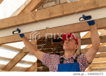 Builder wearing gloves and a red cap is precisely measuring a wooden beam with a spirit level, ensuring accuracy during a construction or renovation project Builder wearing gloves and a red cap is precisely measuring a wooden beam with a spirit level, ensuring accuracy during a construction or renovation project 140956575