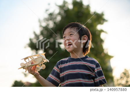 Cheerful Asian boy holding a wooden toy airplane and laughing happily at the park during beautiful golden hour sunset. 140957880