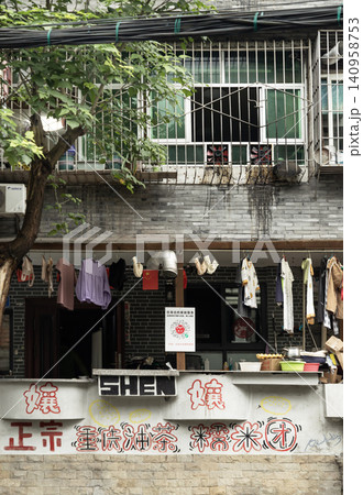 A slice of urban life unfolds against an old grey-brick facade marked by dark rainwater patina. Thick black utility cables stretch horizontally near a leafy tree, while a multi-paned window with A slice of urban life unfolds against an old grey-brick facade marked by dark rainwater patina. Thick black utility cables stretch horizontally near a leafy tree, while a multi-paned window with 140958753