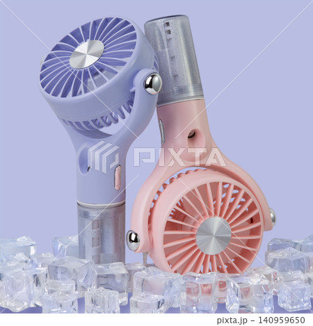 Two pastel pink and purple portable misting fans standing among ice cubes on lavender background, extreme summer cooling concept Two pastel pink and purple portable misting fans standing among ice cubes on lavender background, extreme summer cooling concept 140959650
