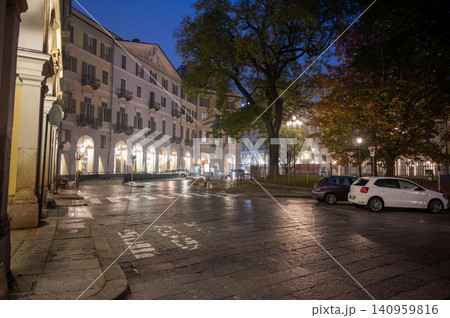 Turin, piedmont, italy. November 20, 2024. Wet cobblestone street reflecting city lights in turin, italy, with historic buildings and trees under a dark evening sky 140959816