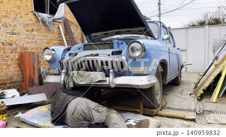 Male repairman lies under his vintage car at courtyard of house. Auto mechanic repairing his blue retro vehicle outdoor. Concept of automobile maintenance. Slow motion Male repairman lies under his vintage car at courtyard of house. Auto mechanic repairing his blue retro vehicle outdoor. Concept of automobile maintenance. Slow motion 140959832