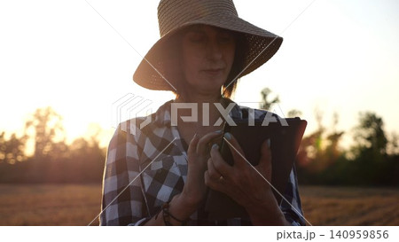 Female agronomist in straw hat monitoring harvest on barley field at sunset. Adult farmer using digital tablet on wheat meadow at dusk. Beautiful scenic landscape. Concept of agricultural business 140959856