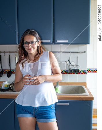 Young woman standing in a modern kitchen, wearing glasses and a white top, holding a glass of water, portraying hydration and a healthy domestic lifestyle Young woman standing in a modern kitchen, wearing glasses and a white top, holding a glass of water, portraying hydration and a healthy domestic lifestyle 140959902