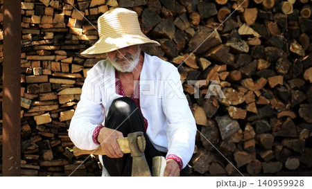 Older man wearing an embroidered shirt gently trims a piece of wood with axe against background of woodshed. Concept of craftsmanship, tradition and ukrainian heritage. Slow motion Older man wearing an embroidered shirt gently trims a piece of wood with axe against background of woodshed. Concept of craftsmanship, tradition and ukrainian heritage. Slow motion 140959928
