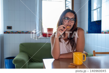 Young woman wearing glasses sitting at a table in the kitchen, eating cookies and drinking from a yellow mug, contemplating during a relaxed morning at home 140960045