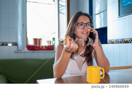 Young woman wearing glasses looking confused and holding a pastry while having a serious conversation on a mobile phone at a kitchen table Young woman wearing glasses looking confused and holding a pastry while having a serious conversation on a mobile phone at a kitchen table 140960049