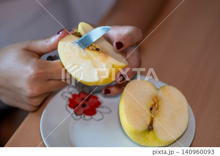 Female hands holding a quartered apple, using a small knife to remove the core and seeds, preparing a healthy fruit snack on a plate with another apple half nearby 140960093