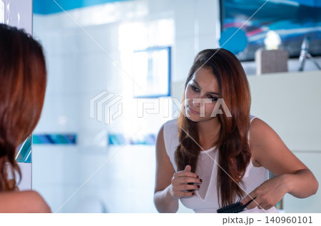 Young woman standing in a bright bathroom, brushing her long, reddish brown hair while looking at her reflection in the mirror, performing a self care routine Young woman standing in a bright bathroom, brushing her long, reddish brown hair while looking at her reflection in the mirror, performing a self care routine 140960101