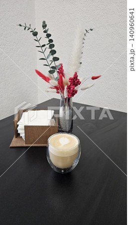 Glass cup of latte on dark table with wooden napkin holder and decorative vase with red and green details in cozy cafe style atmosphere Glass cup of latte on dark table with wooden napkin holder and decorative vase with red and green details in cozy cafe style atmosphere 140960641