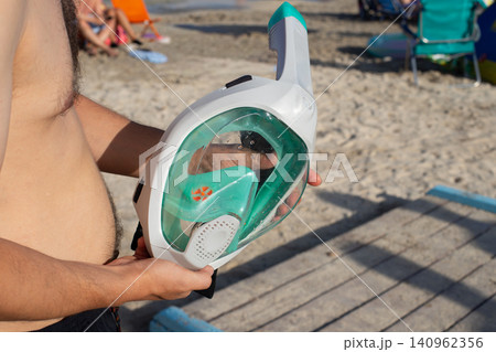 Man holding a full-face snorkel mask while standing on a sandy beach with colorful beach chairs and people in the background Man holding a full-face snorkel mask while standing on a sandy beach with colorful beach chairs and people in the background 140962356