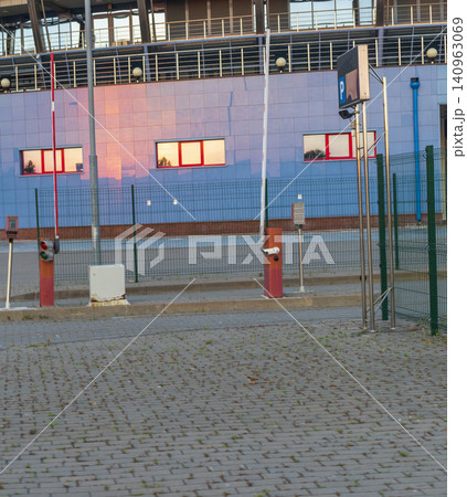 Automated parking lot entrance with barrier gates, security ticket terminal, and a wire mesh safety fence outside a modern stadium building. Automated parking lot entrance with barrier gates, security ticket terminal, and a wire mesh safety fence outside a modern stadium building. 140963069
