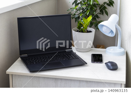Modern home office workspace with a black laptop, smartphone, wireless mouse, blue desk lamp, and green potted plants on a light wooden table. Interior Modern home office workspace with a black laptop, smartphone, wireless mouse, blue desk lamp, and green potted plants on a light wooden table. Interior 140963135