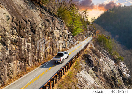 Scenic mountain road in North Carolina Highlands region. Car navigates curves beneath golden trees and steep rocky slopes in fall season. Scenic mountain road in North Carolina Highlands region. Car navigates curves beneath golden trees and steep rocky slopes in fall season. 140964551