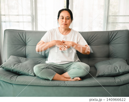 Middle aged Asian woman sitting cross legged on sofa and practicing breathing exercise at home, concept of wellness, meditation, stress relief, mindfulness, and self care. Middle aged Asian woman sitting cross legged on sofa and practicing breathing exercise at home, concept of wellness, meditation, stress relief, mindfulness, and self care. 140966622