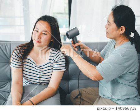 Asian mother drying adult daughter hair with hair dryer at home, concept of family care, personal hygiene, beauty routine, caregiving, bonding, and daily lifestyle. Asian mother drying adult daughter hair with hair dryer at home, concept of family care, personal hygiene, beauty routine, caregiving, bonding, and daily lifestyle. 140966718