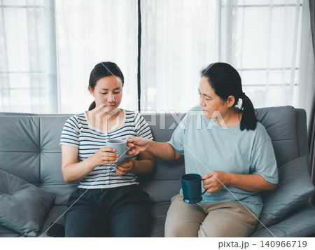 Asian mother giving cup of tea to adult daughter on sofa at home, concept of family support, caregiving, emotional comfort, recovery, kindness, and home wellness. Asian mother giving cup of tea to adult daughter on sofa at home, concept of family support, caregiving, emotional comfort, recovery, kindness, and home wellness. 140966719