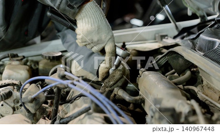 Car mechanic wearing gloves is using a ratchet to skillfully repair the engine of a vehicle with the hood raised, surrounded by tools in a busy garage workshop Car mechanic wearing gloves is using a ratchet to skillfully repair the engine of a vehicle with the hood raised, surrounded by tools in a busy garage workshop 140967448