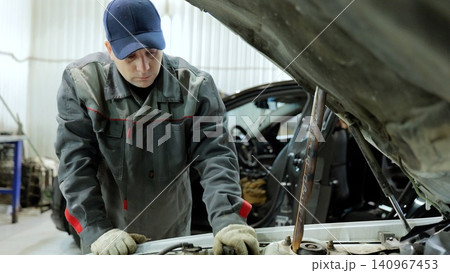 Skilled automotive technician in workshop attire inspecting vehicle engine compartment, checking mechanical components during diagnostic service Skilled automotive technician in workshop attire inspecting vehicle engine compartment, checking mechanical components during diagnostic service 140967453