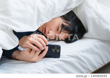 布団にくるまりスマートフォンを操作している若い男性 ライフスタイルイメージ 布団にくるまりスマートフォンを操作している若い男性 ライフスタイルイメージ 140968485