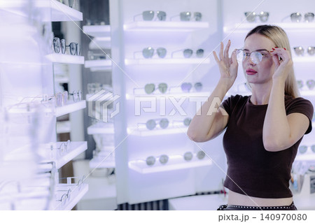 Young woman trying on eyeglasses in optical store, happy customer shopping for new eyewear Young woman trying on eyeglasses in optical store, happy customer shopping for new eyewear 140970080