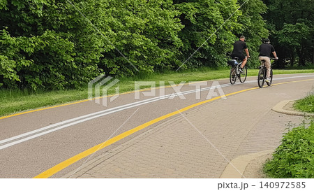 Two cyclists riding away on a marked bike lane through a lush green park Two cyclists riding away on a marked bike lane through a lush green park 140972585