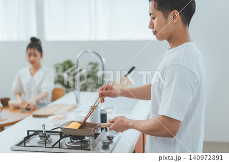 手前でキッチンに立ち玉子焼きを焼く男性と背景で朝ごはんを食べる女性 手前でキッチンに立ち玉子焼きを焼く男性と背景で朝ごはんを食べる女性 140972891