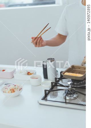 白いキッチンで玉子焼きと弁当の食材が並ぶ弁当作りシーンと菜箸を持つ人物の顔なしショット 白いキッチンで玉子焼きと弁当の食材が並ぶ弁当作りシーンと菜箸を持つ人物の顔なしショット 140972895