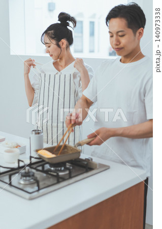 キッチンで男性が玉子焼きを焼く横で女性がエプロンの紐を結ぶカップルの弁当作りシーン キッチンで男性が玉子焼きを焼く横で女性がエプロンの紐を結ぶカップルの弁当作りシーン 140973338