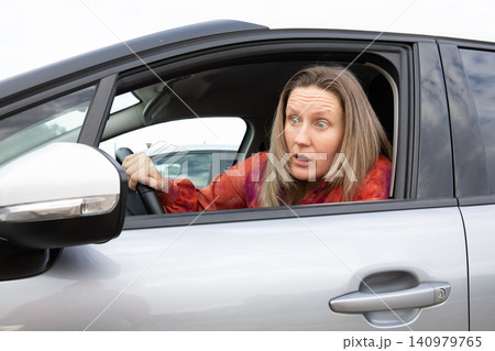 Driver blonde woman looks surprised in car looking outside in parking lot during daytime. Horizontal. 140979765