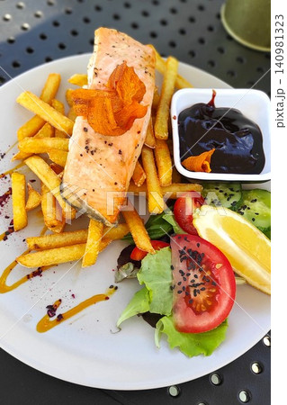 Grilled salmon fillet served with crispy fries, fresh salad, and a side of sauce on a white plate against a textured background 140981323