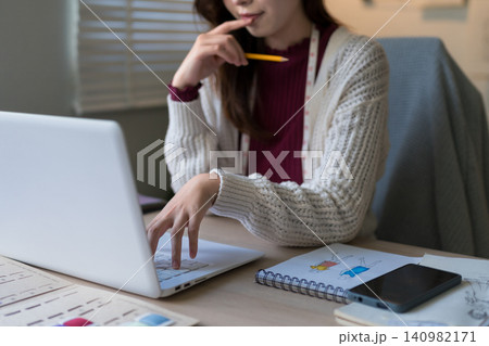 Young Caucasian fashion designer thinking while using laptop reviewing sketches and charts at home office desk Young Caucasian fashion designer thinking while using laptop reviewing sketches and charts at home office desk 140982171